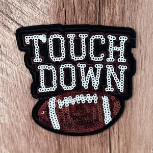 Puede incluir: Un parche decorativo con la palabra TOUCH DOWN en lentejuelas blancas sobre un fondo negro, situado sobre un gráfico de balón de fútbol americano de lentejuelas marrones con cordones y rayas de lentejuelas blancas, bordeado en negro.