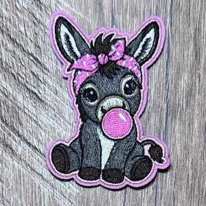 May include: An embroidered iron-on patch featuring a dark gray donkey with a white muzzle, wearing a pink polka-dot bandana on its head. The donkey is blowing a bright pink bubble with bubblegum, outlined in a light purple border.