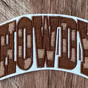 May include: An embroidered patch featuring the word HOWDY in a blocky, arched font. The letters are filled with a checkered pattern alternating between dark brown and light tan thread, all outlined with a thick white border.