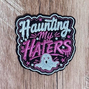 May include: An embroidered patch with a black background featuring the text Haunting My Haters in light blue and pink thread. The design includes a small light blue ghost at the bottom, surrounded by pink and light blue stars and decorative accents.