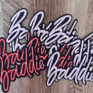 May include: A collection of embroidered patches featuring the word Baddie in a cursive script. The patches have a black glitter background with either light pink or red stitched outlines and detailing, creating a layered and textured appearance.