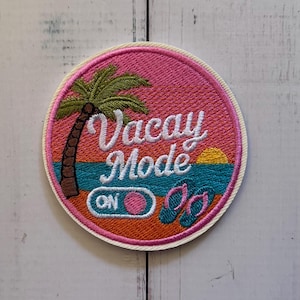 Puede incluir: Un parche bordado circular que presenta un cielo rosa, una palmera verde, un océano azul y un sol amarillo. El texto Vacay Mode está bordado en blanco, con un interruptor etiquetado como ON y un par de chanclas de color turquesa y rosa.
