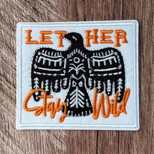May include: A square embroidered patch with a white background and a black bird silhouette featuring white geometric patterns. The text LET HER is stitched in orange above the bird, and the text Stay Wild is stitched in orange below the bird.