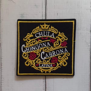 May include: A square black embroidered patch featuring a gold filigree border and red rose accents. The text CHULA, CHINGONA, CABRONA, and LATINA is stitched in white thread across the center, arranged within the ornate gold decorative frame.