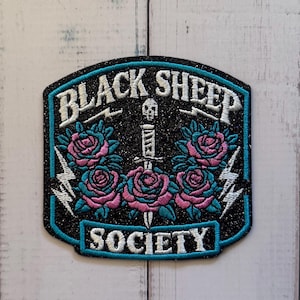 May include: A black glittery embroidered patch featuring the text BLACK SHEEP SOCIETY in white. The design includes a central dagger with a skull pommel, surrounded by pink and teal roses, and white lightning bolts, all bordered by a teal line.