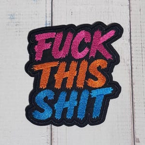 May include: A black embroidered patch featuring the text FUCK THIS SHIT in three distinct colours. The word FUCK is in pink, THIS is in orange, and SHIT is in blue. The patch has a black border and a textured, stitched appearance throughout.