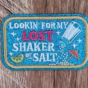 Könnte beinhalten: Ein rechteckiger bestickter Aufnäher mit blauem Glitzerhintergrund und goldenem Rand. Er zeigt den Text LOOKIN' FOR MY LOST SHAKER OF SALT in Weiß und Pink sowie eine Abbildung eines Salzstreuers, einer Limettenscheibe und eines Cocktailschirms.