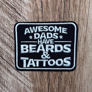 May include: A rectangular black embroidered patch with a white border and white stitched text that reads AWESOME DADS HAVE BEARDS & TATTOOS, featuring two small stars flanking the word DADS and a horizontal line separating the text lines.