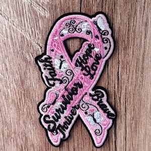 Könnte beinhalten: Ein rosa Awareness-Schleifen-Aufnäher mit schwarzem Stickrand und schwarzem Text mit den Wörtern Faith, Hope, Love, Survivor und Brave. Das Design zeigt filigrane schwarze Schnörkelmuster und kleine weiße Schmetterlingsakzente auf dem rosa Stoff.