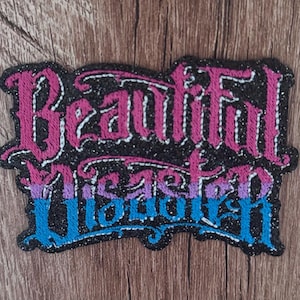 May include: An embroidered patch with a black glitter background featuring the text Beautiful Disaster. The word Beautiful is stitched in pink, while the word Disaster features a gradient transition from purple to blue, all in a gothic font.