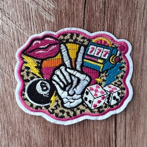 May include: An embroidered patch featuring a central skeleton hand making a peace sign, surrounded by a leopard print background, a pink pair of lips, a blue slot machine with 777, two white dice with pink dots, and a black eight ball.