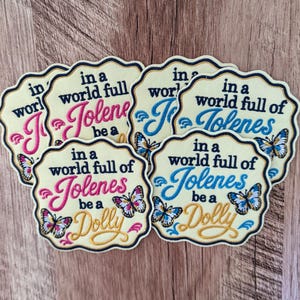 May include: A collection of scalloped-edge embroidered patches on a light cream background. Each patch features the black text "in a world full of Jolenes be a Dolly" with colorful butterflies and either pink or blue embroidered accents.