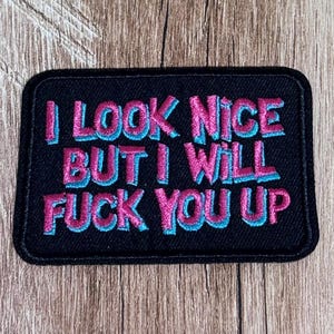 May include: A rectangular black embroidered patch with rounded corners featuring the text I LOOK NICE BUT I WILL FUCK YOU UP in bold, blocky letters. The lettering is stitched in a vibrant pink colour with a light blue shadow effect.