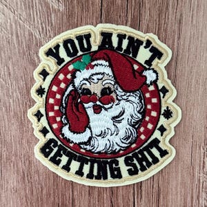May include: A circular embroidered patch featuring a Santa Claus figure with a white beard, red hat, and red gloves. The background is a red and white checkered circle. The text "YOU AIN'T GETTING SHIT" is embroidered in black around the border.