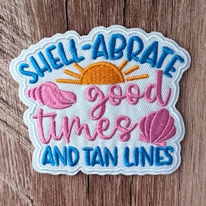 Pode incluir: Um emblema bordado branco com borda recortada apresentando um sol amarelo, duas conchas rosas e texto em azul e rosa que diz: SHELL-ABRATE good times AND TAN LINES. O design está centralizado em um fundo de tecido branco.