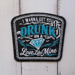 May include: A black embroidered patch with a glittery finish featuring the text I WANNA GET YOU DRUNK ON A LOVE LIKE MINE in white and teal thread. The design includes a central diamond graphic outlined in white and teal accents.