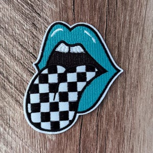 May include: An embroidered patch shaped like a pair of open lips with teal colored edges, white highlights, and a black interior. A large tongue protrudes from the mouth, featuring a black and white checkered pattern with a white border.