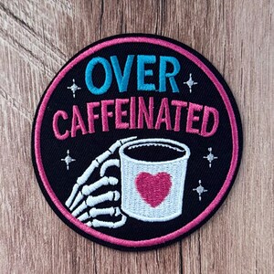May include: A circular embroidered patch with a black background and a pink border. It features the text OVER CAFFEINATED in blue and pink, a white skeleton hand holding a white mug with a pink heart, and small white star accents.