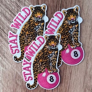May include: Three embroidered patches featuring a leopard sitting on a pink billiard ball with the number eight. The text "STAY WILD" is stitched in pink thread along the side of each patch. The leopard has black spots on a golden brown coat.