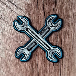 May include: An embroidered patch featuring two crossed silver wrenches with a bright blue border and a black outline. The design is rendered in a detailed stitch pattern, showcasing the metallic texture of the tools in a clean, graphic style.