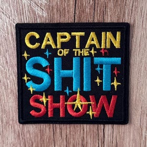 May include: A square black embroidered patch featuring the yellow text CAPTAIN OF THE, the bright blue text SHIT, and the red text SHOW. The design is accented with small, colorful embroidered star shapes in yellow, blue, and red throughout.