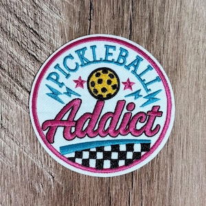 May include: A circular embroidered patch with a white background and a hot pink border. It features the text "PICKLEBALL" in teal, "Addict" in hot pink, a yellow pickleball with black dots, two small pink stars, teal lightning bolts, and a black and white chequered pattern at the bottom.