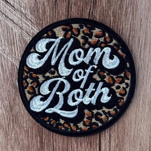 Może przedstawiać: Okrągła haftowana naszywka z tłem w panterkę w odcieniach brązu, beżu i czerni. Napis "Mom of Both" jest wyhaftowany białą, kursywną czcionką w stylu retro na środku, otoczony grubą czarną obwódką.