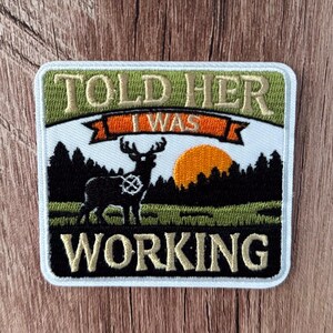 May include: A square embroidered patch featuring a black silhouette of a deer standing in a field before a forest and an orange sun. The text TOLD HER I WAS WORKING is stitched in beige thread with an orange banner accenting the middle section.