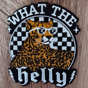 May include: An embroidered patch featuring a leopard with white sunglasses against a black and white checkered background. The text WHAT THE is arched above the leopard, and the word helly is written below, flanked by two white lightning bolt icons.