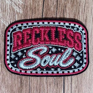May include: An oval embroidered patch with a black background featuring the text RECKLESS in red block letters and Soul in silver script. The design includes small silver star accents and a checkered border in black and silver thread.