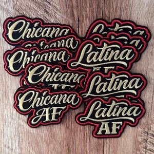 Könnte beinhalten: Eine Sammlung von gestickten Stoffaufnähern mit den Wörtern Chicana, Chicana AF, Latina und Latina AF. Jeder Aufnäher hat goldfarbene Schreibschrift auf schwarzem Hintergrund, umrandet von einem dicken roten Rand in gewellter Form.