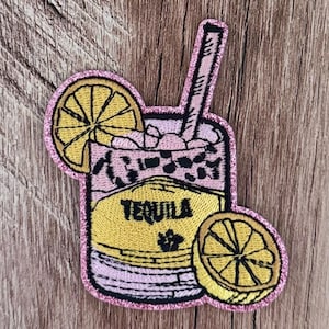 May include: An embroidered patch featuring a cocktail glass with a pink liquid, ice cubes, and a straw. The glass has a yellow label with the word TEQUILA printed in black. Two yellow lemon slices are attached to the glass, and the entire patch is outlined with a shimmering pink glitter border.