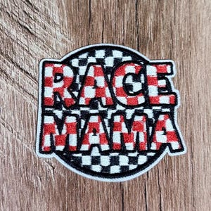 Könnte beinhalten: Ein runder, bestickter Aufnäher mit schwarz-weiß kariertem Hintergrund und dem Text RACE MAMA in fetten, rot-weiß karierten Blockbuchstaben. Der Aufnäher hat einen dicken weißen Rand und ist für die Anbringung auf Textilien vorgesehen.