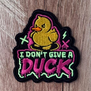 Peut inclure: Un écusson brodé représentant un canard jaune au bec orange faisant un geste grossier. Le motif inclut des accents roses et vert clair avec le texte I DON'T GIVE A DUCK dans une police dégoulinante sur un fond à bordure noire.