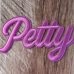 Puede incluir: Un parche bordado de color morado que presenta la palabra Petty en una fuente cursiva. Las letras están cosidas con un hilo denso y brillante, creando una textura en relieve con un borde distintivo que perfila cada letra con claridad.