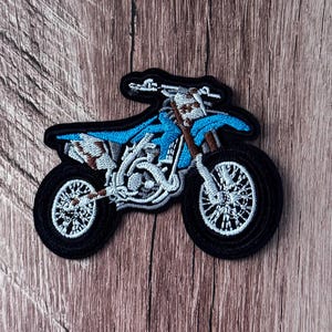 Puede incluir: Un parche bordado con forma de moto de cross. La motocicleta presenta un chasis y asiento de color azul brillante, neumáticos negros con radios blancos y detalles en marrón y blanco en el motor, el manillar y la parte trasera, todo bordeado en hilo negro.