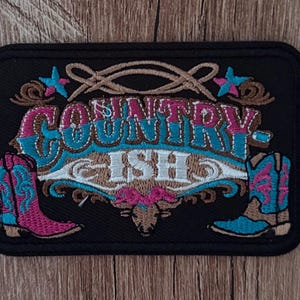May include: A rectangular black embroidered patch featuring the text "COUNTRY-ISH" in white, pink, and blue thread. The design includes two cowboy boots in pink and blue, decorative swirls, and two small blue stars on a black fabric background.
