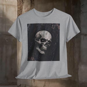 May include: A gray t-shirt featuring a detailed illustration of a skull with dark eye sockets and a detailed jawline. The skull is set against a dark, ornate background. The t-shirt is made of a soft material.