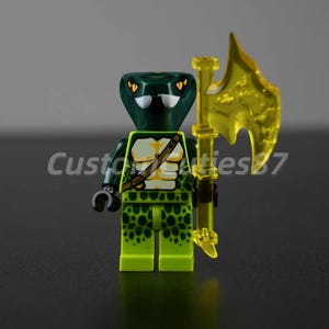 May include: A custom Lego minifigure of a snake-headed warrior. The figure is primarily green with yellow accents, including a translucent yellow axe. The torso features a tan and white design. The text "Customties87" is visible on the left side.