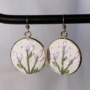 May include: A pair of circular drop earrings featuring gold-toned metal frames and white fabric inserts. Each earring displays hand-stitched embroidery of light purple flower buds with green stems and leaves, suspended from gold-toned metal hooks.