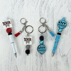 May include: A collection of graduation-themed pens and keychains. The pens are white and blue, with decorative beads and the text "SENIOR" or "GRADUATED." The keychains feature tassels and the same text and graphics.