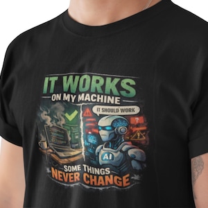May include: Black t-shirt with a colorful graphic featuring a robot and the text "IT WORKS ON MY MACHINE" and "SOME THINGS NEVER CHANGE". The design includes a laptop, check mark, and the phrase "IT SHOULD WORK".