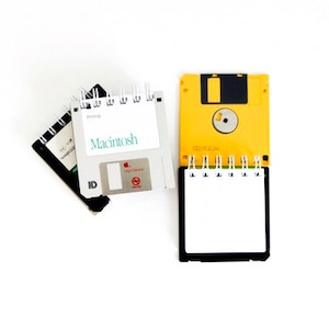 May include: A yellow and black floppy disk notebook with a white page and a black spiral binding. The notebook is open to a blank page. The floppy disk is labeled "3.5" and has a small red apple logo. The notebook is sitting on a white surface.