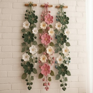 Può includere: Tre decorazioni da parete verticali con fiori e foglie all'uncinetto dettagliati, appesi a bastoncini di legno. Le decorazioni laterali mostrano fiori verde scuro e bianchi, mentre quella centrale presenta fiori all'uncinetto rosa e bianchi.