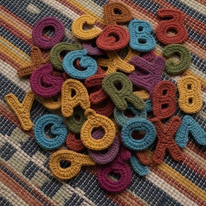 May include: A collection of small, individual letters crocheted from yarn in various colors including mustard yellow, teal, olive green, rust orange, and deep purple. Each letter is crafted with a textured stitch pattern and arranged in a pile.