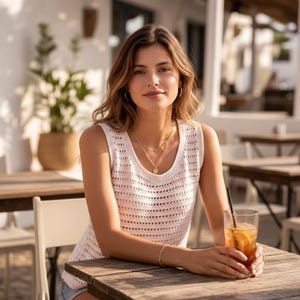 Peut inclure: Une femme porte un débardeur blanc en maille ajourée et des colliers dorés, assise à une table en bois. Elle tient un verre de thé glacé avec une paille. L'arrière-plan comprend un bâtiment blanc, des plantes et des sièges extérieurs.