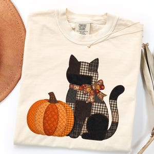 Può includere: Una t-shirt a maniche corte color crema con etichetta Comfort Colors sul collo. Il davanti presenta una grafica in stile patchwork di un gatto nero seduto accanto a una zucca arancione. Il gatto indossa un fiocco fantasia intorno al collo.