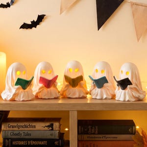 May include: Five white ghost figurines with glowing yellow eyes, each holding a small open book in a different color: green, pink, yellow, blue, and black. The ghosts are arranged in a row on a wooden shelf, illuminated from within.