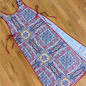 May include: A sleeveless apron with a vibrant, geometric pattern in red, white, blue, and yellow. The apron has red trim and ties, and is laid out on a wooden surface.