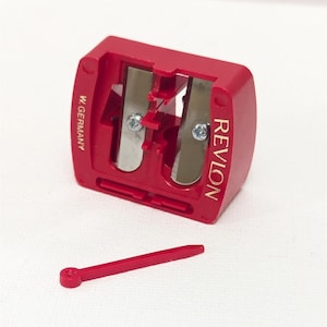 May include: A red Revlon pencil sharpener with two silver blades and a small red cleaning stick. The sharpener is marked "W. Germany" and "Revlon" in gold lettering. The sharpener is designed to sharpen pencils.